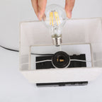 A hand holding a clear LED filament light bulb above a white fabric lampshade with a black socket base, shown against a plain white background.