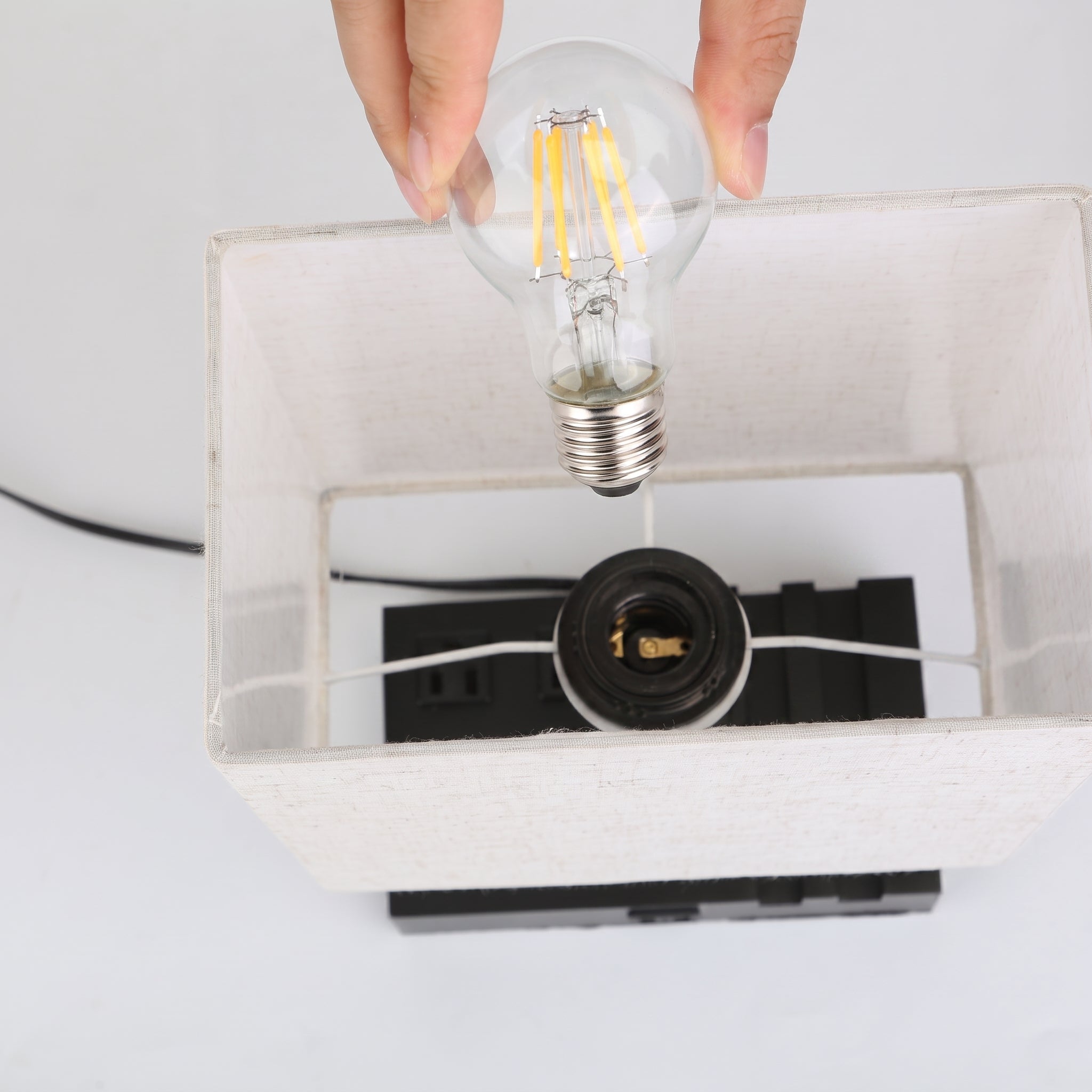 A hand holding a clear LED filament light bulb above a white fabric lampshade with a black socket base, shown against a plain white background.