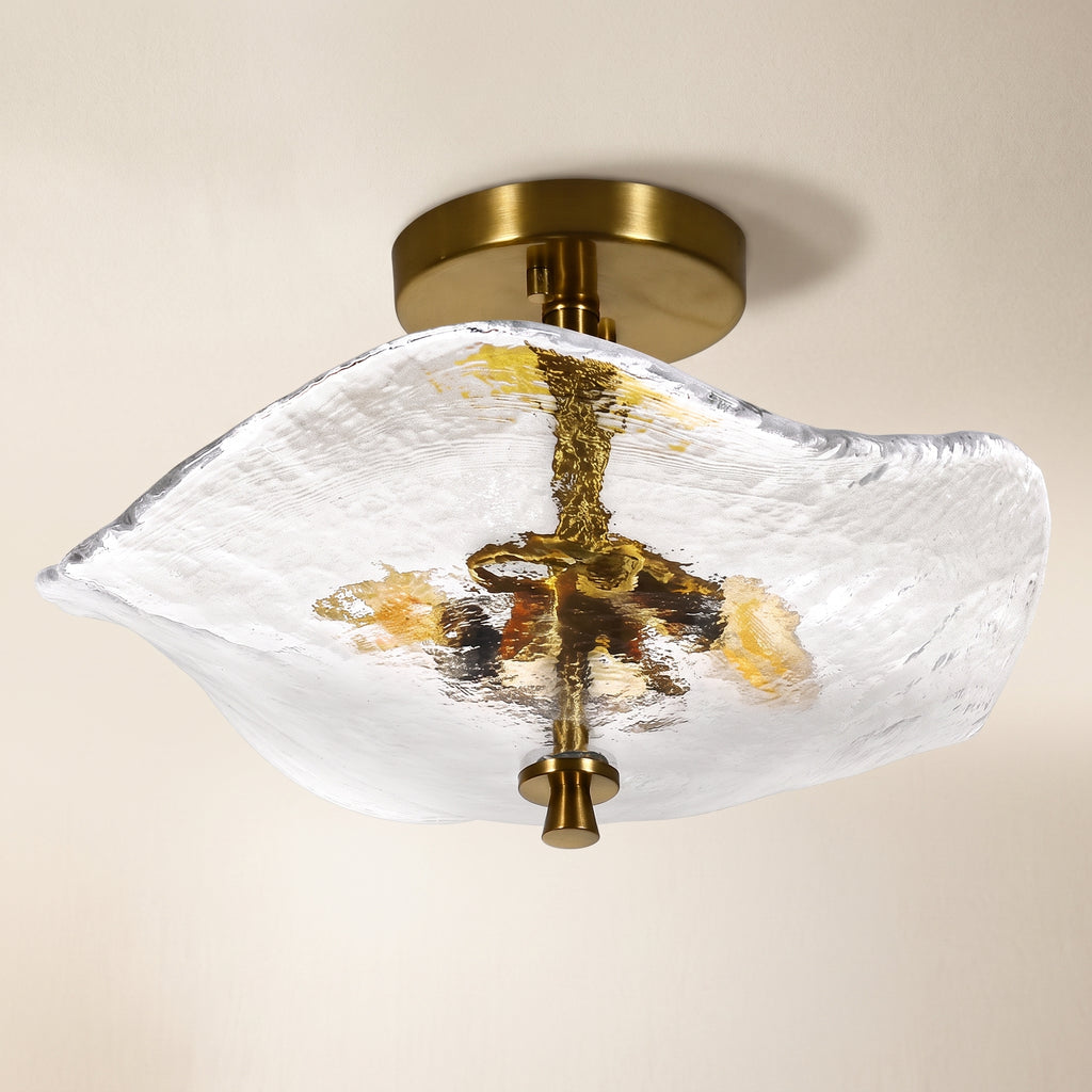 A modern semi-flush mount ceiling light fixture featuring a textured, organic-shaped clear glass shade with gold and amber accents, suspended from a brushed brass ceiling canopy against a warm off-white background.