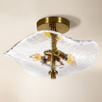 A modern semi-flush mount ceiling light fixture featuring a textured, organic-shaped clear glass shade with gold and amber accents, suspended from a brushed brass ceiling canopy against a warm off-white background.