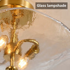 Close-up of a gold metal ceiling light fixture with a textured, wavy clear glass shade, showing illuminated bulbs inside. A text overlay in the top right reads 'Glass lampshade'.