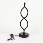 A modern black LED table lamp with a sculptural, twisting infinity-loop design on a round base, shown with its USB charging cable on a white background.