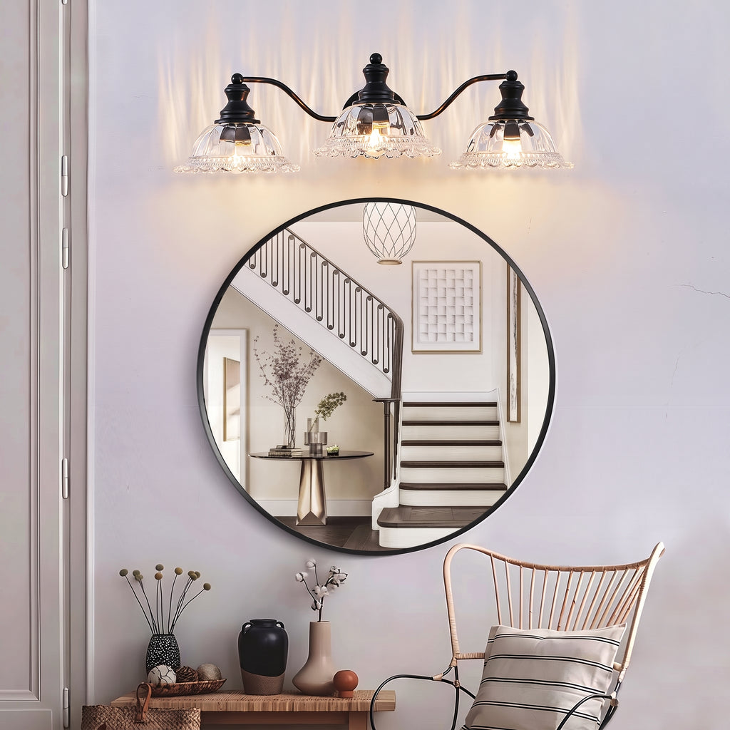 A round wall mirror with a thin black frame hangs on a light gray wall beneath a three-light sconce. The mirror reflects a staircase, and the scene includes a chair and small tabletop decor below.