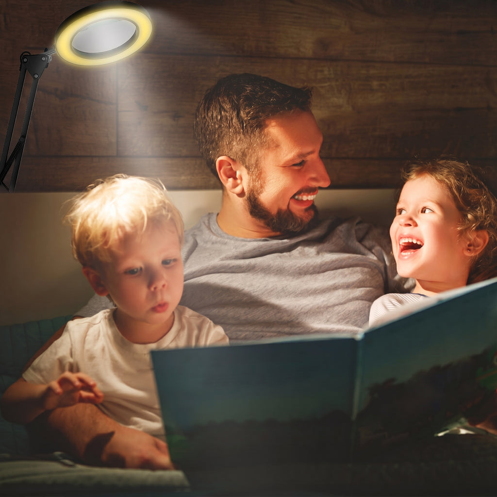 A father and two children are gathered under a wooden ceiling, reading a book illuminated by a glowing ring light attached to a black metal arm. The warm light casts a soft glow on the family's faces and the book.