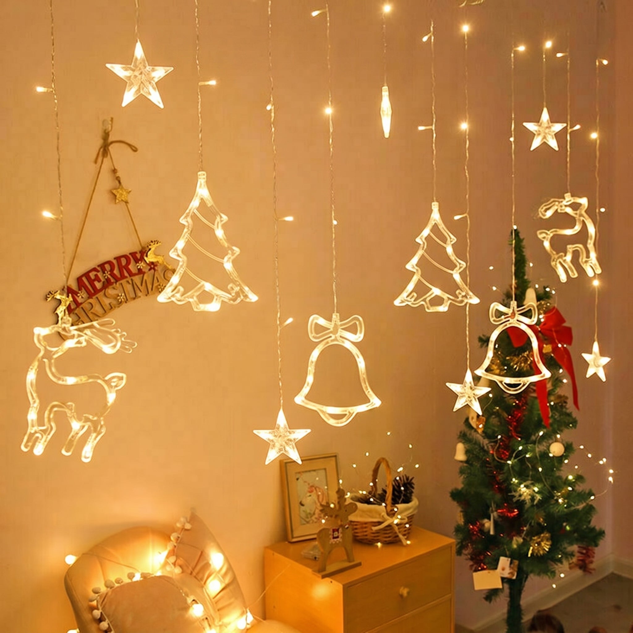 Warm white LED curtain string lights with hanging Christmas ornaments, including stars, bells, trees, and reindeer cutouts, displayed in a cozy decorated room next to a Christmas tree.