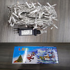 Coiled white LED Christmas string lights with a black power adapter placed on a gray wood-grain surface, with the product box below showing a colorful winter holiday scene and the word LED.