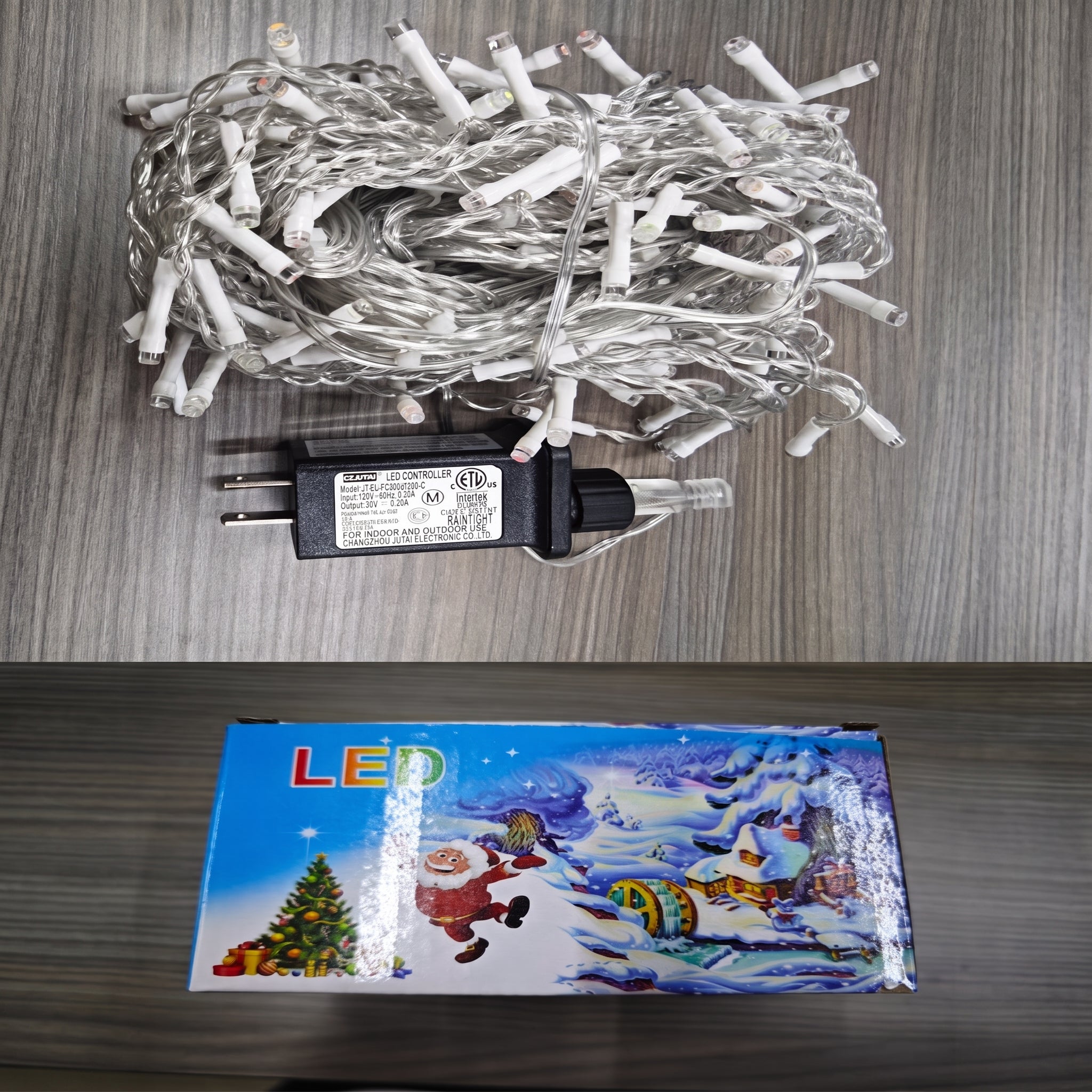 Coiled white LED Christmas string lights with a black power adapter placed on a gray wood-grain surface, with the product box below showing a colorful winter holiday scene and the word LED.