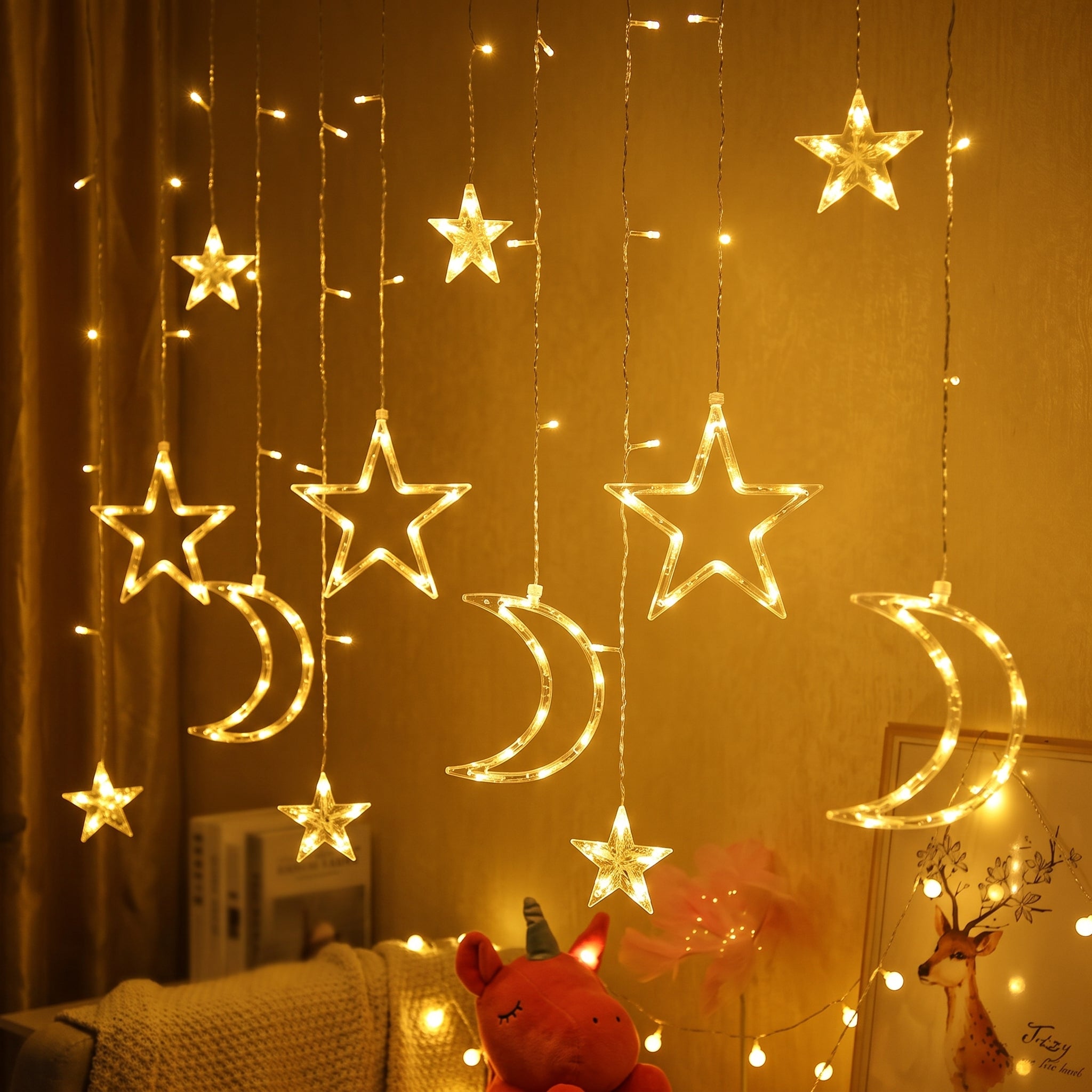 Warm white LED curtain string lights featuring hanging star and crescent moon shapes, displayed in a cozy room setting with a knitted blanket and stuffed animal.